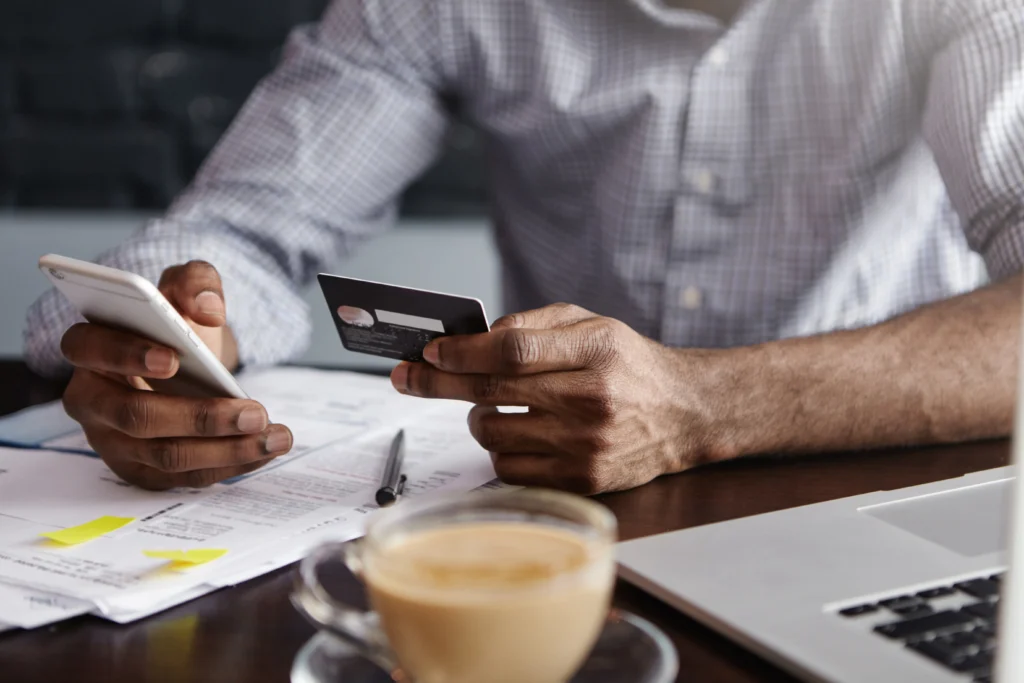 Homem cadastrando cartão de crédito no celular com notebook e café, mostrando praticidade no uso de cartões sem anuidade