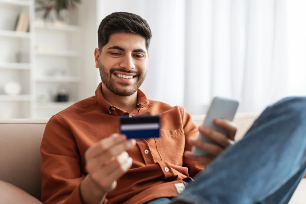 Homem no sofá olhando para seu cartão de crédito com celular na mão, simulando controle financeiro e uso consciente.