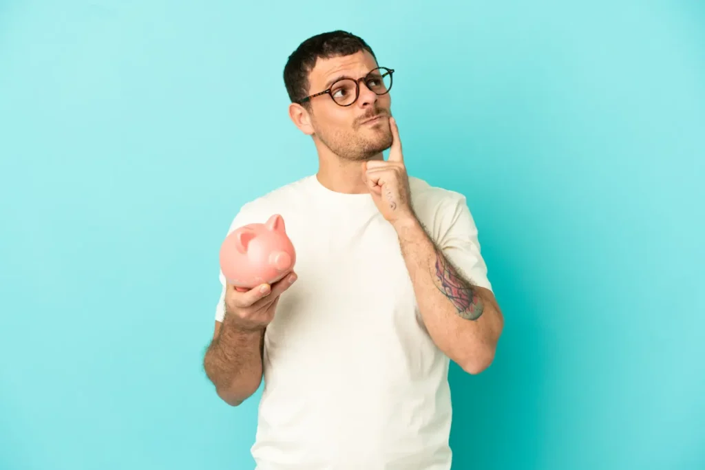 Homem brasileiro tatuado pensativo segurando um cofrinho rosa, refletindo sobre como a consultoria financeira ajuda a criar reserva de emergência