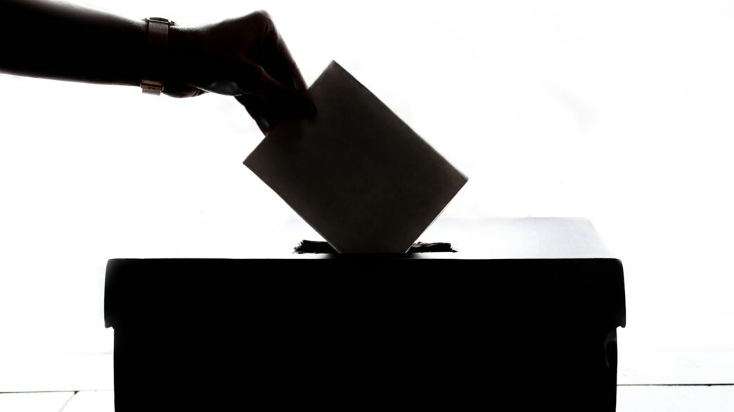 Pessoa depositando o voto em uma urna, em fundo escuro